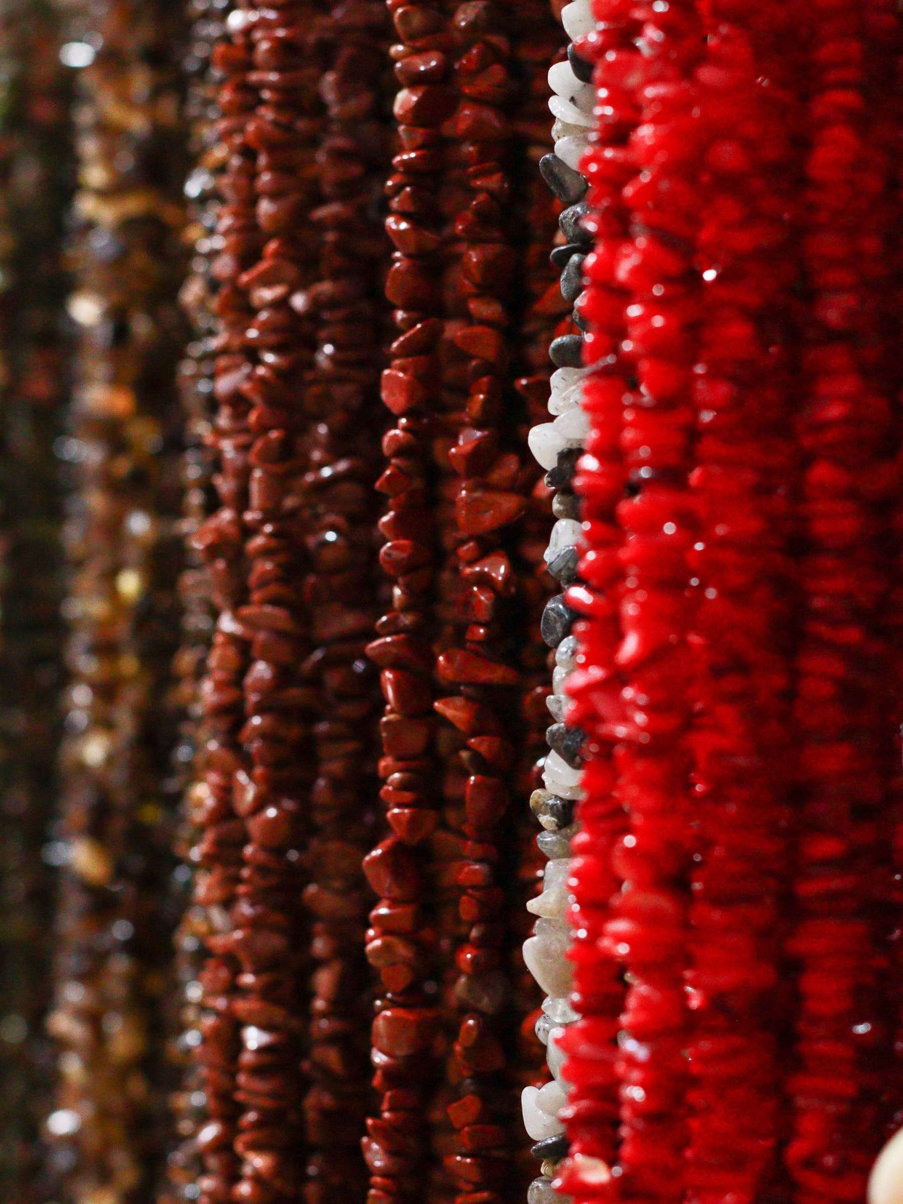 Fios de pedras em tons de vermelho, coral e marrom pendurados lado a lado na Kita Centro